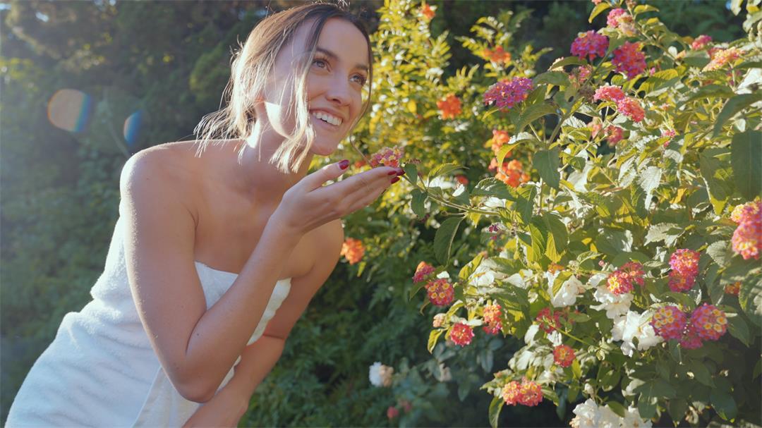 Beautiful girl outdoors with white bath towel around a sunny day admiring flowers and smelling flowers